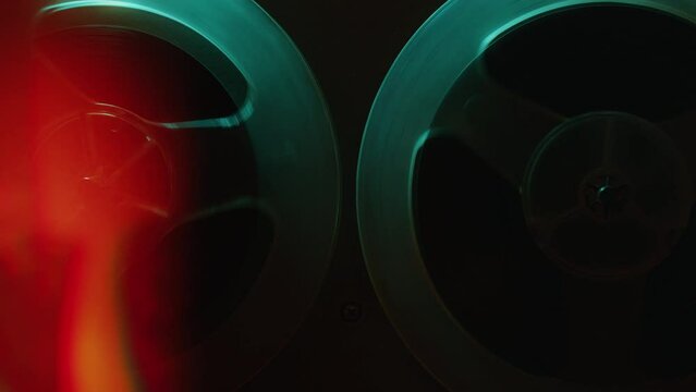 Retro-styled open reel tape recorder with rotating spools in dark studio with blue neon light. Zoom off, close-up shot