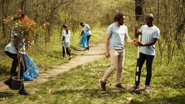 Team of climate change activists planting green trees and seeds in a forest environment, uniting to conserve ecosystem. Volunteers installing plants in the ground, nurture greenery. Camera B.