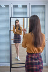 Office girl wearing a plaid mini skirt stands looking at her reflection in a full-length mirror in her dressing room at home.