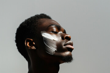 Close-up of a young man with a thoughtful expression, a dab of moisturizing cream on his cheek, highlighting a daily skincare routine.