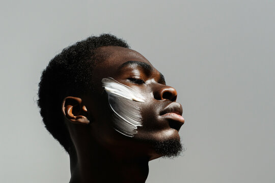 Close-up of a young man with a thoughtful expression, a dab of moisturizing cream on his cheek, highlighting a daily skincare routine.