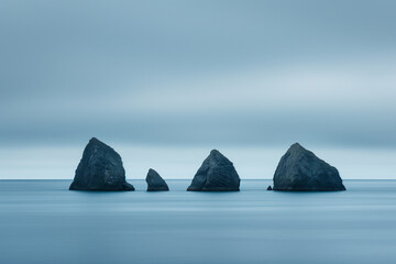 Four solemn sea stacks emerge from the depths of a calm ocean, set against a soft, diffused light...