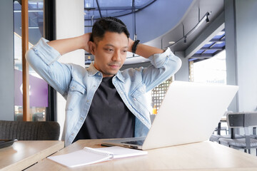 Asian man Tired and exhausted with blue shirt using laptop and mobile phone in coffee shop, online freelance business