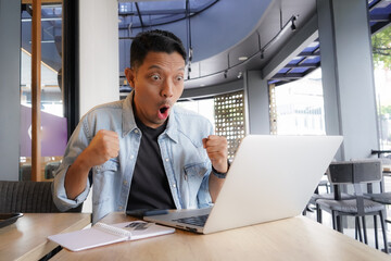 Asian man shocked face, wow, surprised with blue shirt using laptop and mobile phone in coffee...