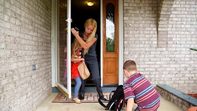 A mother is trying to leave the house talking on a phone with 2 children who are unprepared and dropping their book bags for school for a family stress, schedule or career concept.