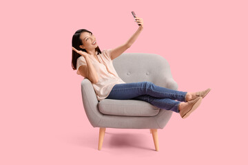 Beautiful Asian woman with mobile phone taking selfie in grey armchair on pink background - obrazy, fototapety, plakaty