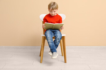 Fototapeta premium Little boy reading book while sitting on chair near beige wall