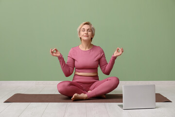 Mature woman with laptop meditating while sitting near green wall. Online yoga classes