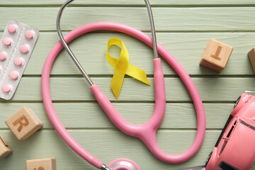 Golden ribbon with stethoscope, pills and toys on green wooden background. Children cancer...