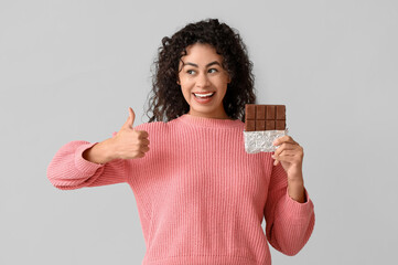 Happy young African-American woman with sweet chocolate bar showing thumb-up gesture on grey...