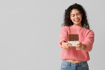 Happy young African-American woman with sweet chocolate bar on grey background