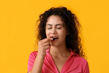 Beautiful young African-American woman eating sweet chocolate candy on yellow background