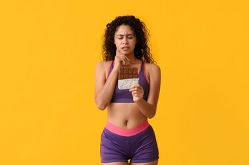 Thoughtful young African-American woman with sweet chocolate bar on yellow background - obrazy, fototapety, plakaty