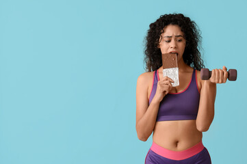 Sporty young African-American woman with sweet chocolate bar and dumbbell on blue background