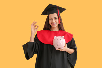 Happy female graduate with piggy bank showing OK gesture on yellow background. Tuition fees concept