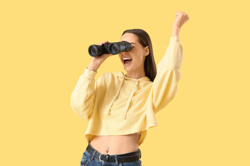 Happy young woman looking through binoculars on yellow background