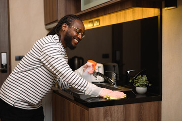 Happy African man wearing protective glove cleaning the kitchen counter with cleaning spray and cloth - Powered by Adobe