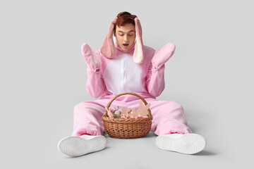 Surprised young man in Easter bunny costume sitting with basket on white background