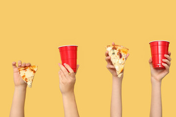 Many hands holding tasty pizza slices and one-use cups on yellow background