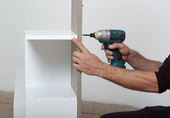 Man assembling a white shelf. A power drill is being used to attach a wooden plank to the shelf. The mans hands are visible, indicating the process of assembly. The shelf is against a white wall.