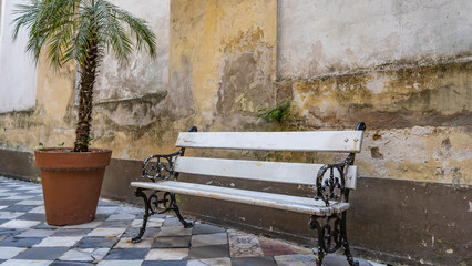 An old wooden white bench with openwork curved metal legs stands against a weathered wall. The...