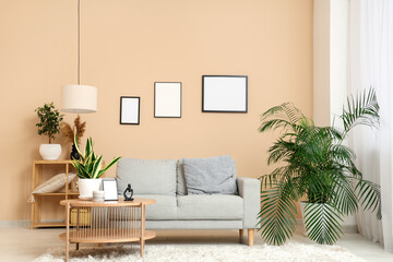 Stylish living room with cozy sofa, houseplants and blank picture frames on beige wall - obrazy, fototapety, plakaty