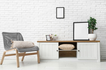 Armchair and chest of drawers with houseplant and blank picture frames near white brick wall in room