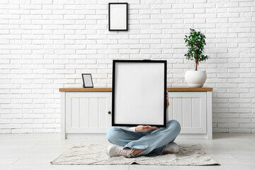 Young woman sitting near chest of drawers and holding blank picture frame in room - Powered by Adobe
