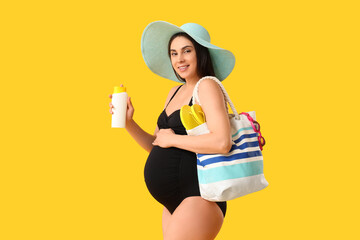 Young pregnant woman in swimsuit and hat holding bag and sunscreen on yellow background. Travel...