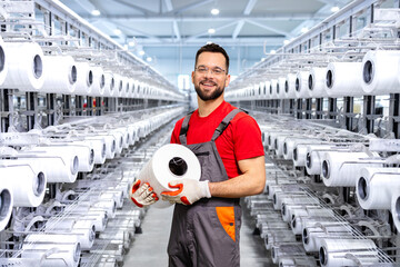 Portrait of experienced worker in textile factory holding thread spool and standing by industrial knitting machine.