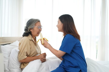 Asian young caregiver female feeding banana aged woman on bed at home