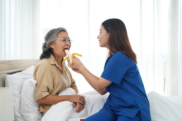 Asian young caregiver female feeding banana aged woman on bed at home