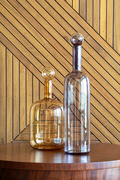 Two transparent glass decanters with ornate stoppers resting on a polished wooden surface, complemented by a herringbone-patterned wooden backdrop