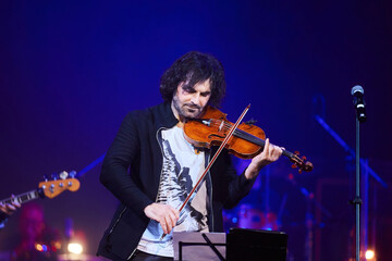 A violinist musician stands on stage with a violin in his hands during a concert. There is a bright light from the floodlights around. - 789874619