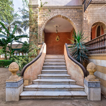 White marble staircase with ornate stone railings, flanked by sculptures, that descends to a serene courtyard surrounded by lush greenery and tropical plants