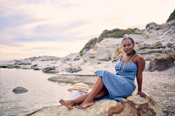 Portrait, beach or black woman on rock to relax on holiday vacation or resting break in Greece at sunset. Tourist, girl or African person sitting at ocean, nature or sea for peace, wellness or travel