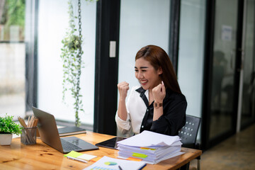 Happy Asian businesswoman raising her fists in excitement at a job well done at her desk in a modern office.