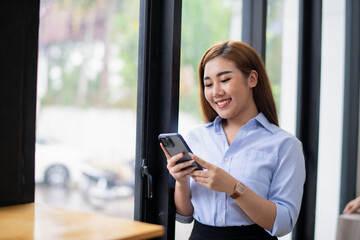 Fototapeta na wymiar Smiling young Asian businesswoman using her smartphone by the window in a well-lit office, dressed in smart casual attire.