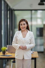 Portrait of a poised Asian businesswoman in a white suit standing confidently in a contemporary office workspace.