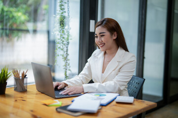 Professional Asian businesswoman typing on laptop at a wooden desk with paperwork and greenery in a...