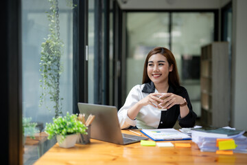 Professional Asian businesswoman typing on laptop at a wooden desk with paperwork and greenery in a bright office setting.