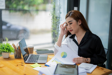 Fototapeta premium Professional Asian businesswoman typing on laptop at a wooden desk with paperwork and greenery in a bright office setting.