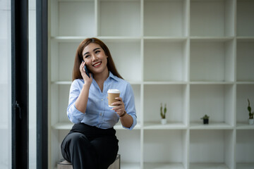 Fototapeta premium Smiling young Asian businesswoman using her smartphone by the window in a well-lit office, dressed in smart casual attire.