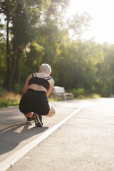 A fit young woman with short hair prepares to run, crouched on a sunlit path with trees in the...