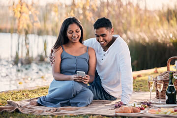 Couple, picnic and phone for social media in outdoors, nature and relax on grass for bonding. People, lake and app for funny joke or online humor on vacation, travel and browse tech on park date
