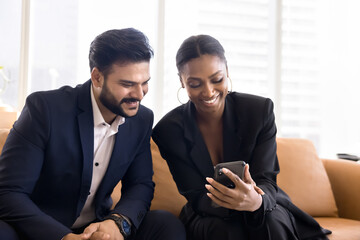 Cheerful multiethnic office friends in formal suits watching funny online content on smartphone together, sitting on couch, using mobile phone, looking at screen, laughing, enjoying communication