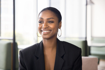 Happy beautiful young African professional woman in formal jacket posing in co-working office, looking away with toothy smile. Confident businesswoman, manager, legal expert portrait.