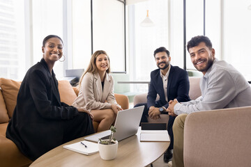 Multiethnic team of happy coworkers meeting in comfortable co-working space, sitting on couch, in armchairs, looking at camera, smiling, posing for business group portrait, promoting diversity