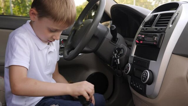 A little boy in a white T-shirt sits behind the wheel in a car and shifts gears on the gearbox. The child is playing in Dad's car.