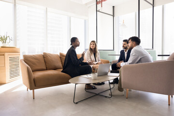 Young African American professional woman talking to diverse team in comfortable office co-working space, sitting on couch, speaking to multiethnic colleagues at corporate meeting